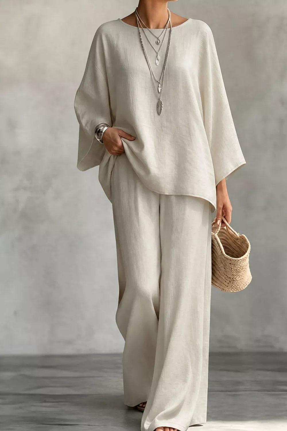 NEWYORKLISTED STYLE COLLECTION: A woman wearing a loose, two-piece linen outfit in a warm off-white or cream color, consisting of a wide-sleeve top and matching wide-leg pants. She is accessorized with layered silver necklaces, a silver watch, and holds a small woven straw handbag against a textured gray studio background.