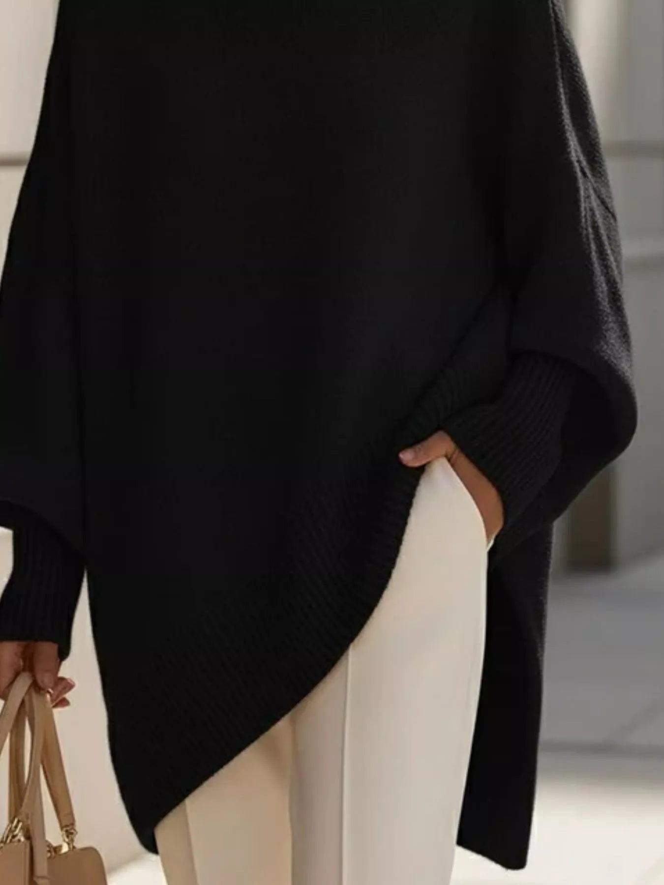 NEWYORKLISTED STYLE COLLECTION: Person wearing a black poncho and white pants, holding a beige handbag indoors.