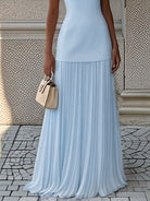 NEWYORKLISTED STYLE COLLECTION: Person wearing a light blue pleated dress holding a beige handbag against a textured wall.