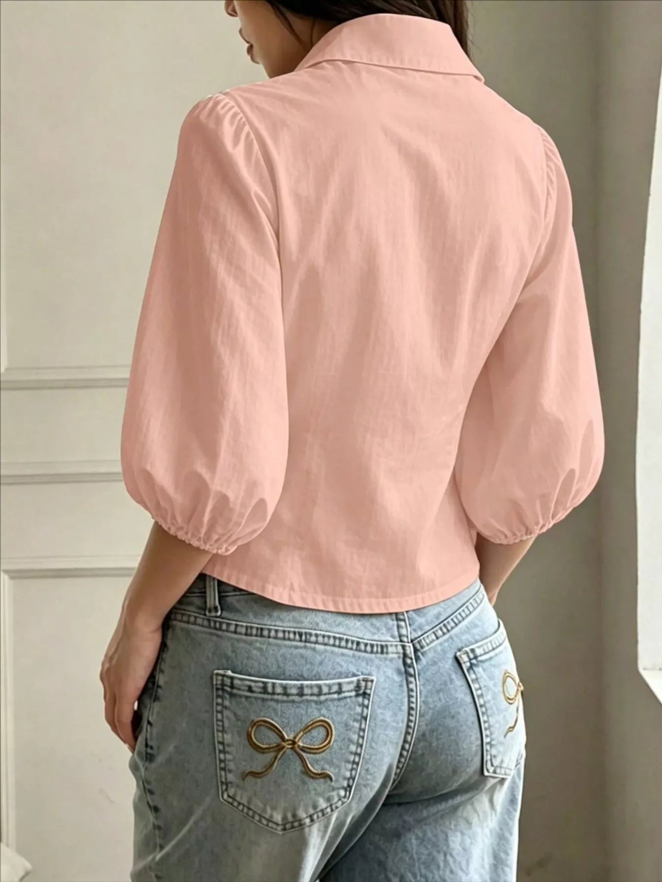 NEWYORKLISTED STYLE COLLECTION: A woman shown from the back wearing a light pink cropped blouse with voluminous elbow-length puff sleeves and light-wash blue jeans with decorative bow embroidery on the back pockets.