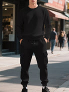 NEWYORKLISTED ATLAS MEN'S COLLECTION: A man wearing a black textured crewneck sweatshirt and matching black cargo jogger pants stands on a city sidewalk with blurred storefronts and pedestrians in the background.