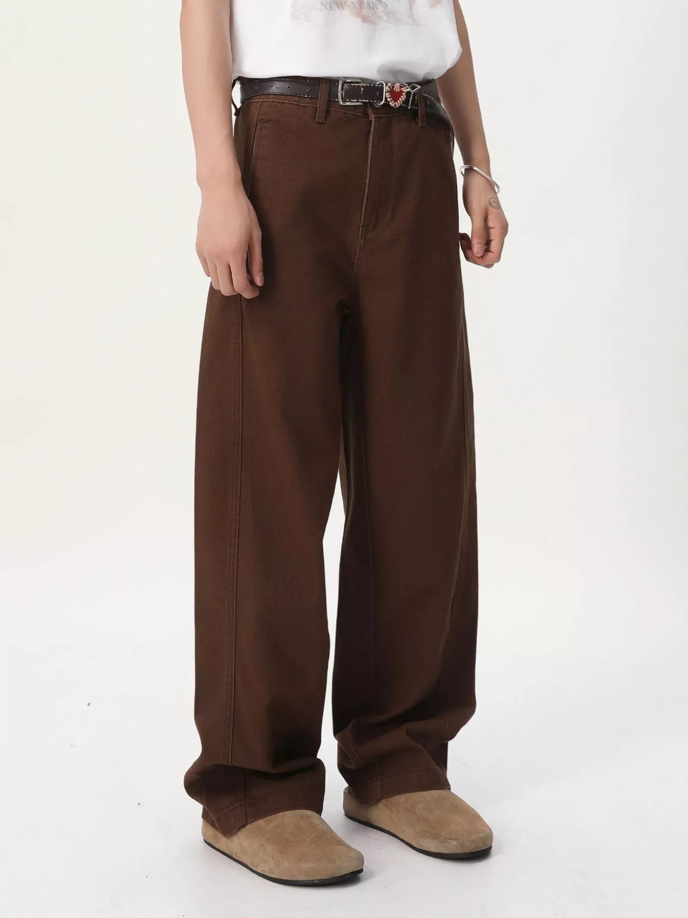 NEWYORKLISTED ATLAS MEN'S COLLECTION: A person wearing loose, wide-leg brown pants with a black belt, a white top, and tan slip-on shoes, photographed against a plain light background.