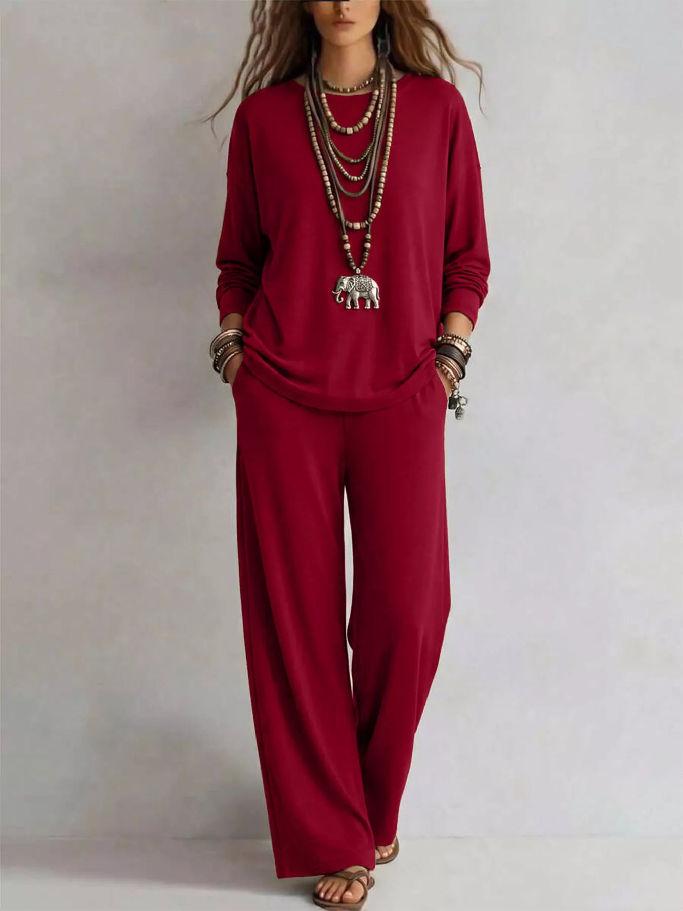 NEWYORKLISTED STYLE COLLECTION: A woman modeling a matching deep red long-sleeve top and wide-leg pants set, accessorized with layered silver necklaces, bracelets, and sandals, against a plain light gray background.