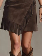 NEWYORKLISTED STYLE COLLECTION: A close-up of a model wearing a brown suede fringe mini skirt and matching brown cowboy boots against a neutral gray background.