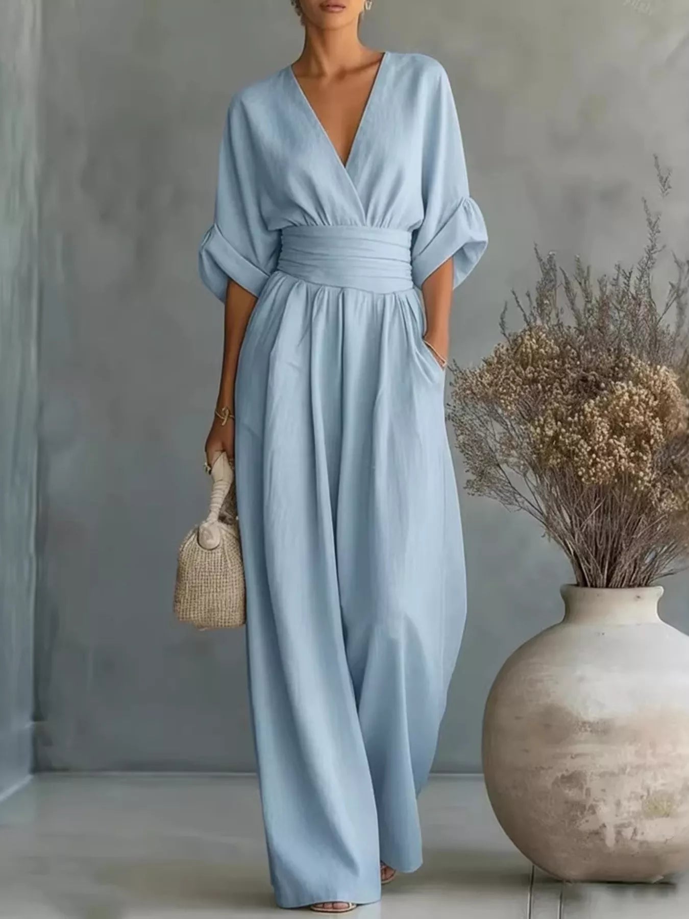 NEWYORKLISTED STYLE COLLECTION: Woman wearing a light blue dress standing next to a vase with dried flowers against a gray wall.