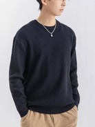 NEWYORKLISTED ATLAS MEN'S COLLECTION: A man wearing a dark navy ribbed knit crewneck sweater, paired with beige pants and a silver necklace, photographed against a light gray studio background.