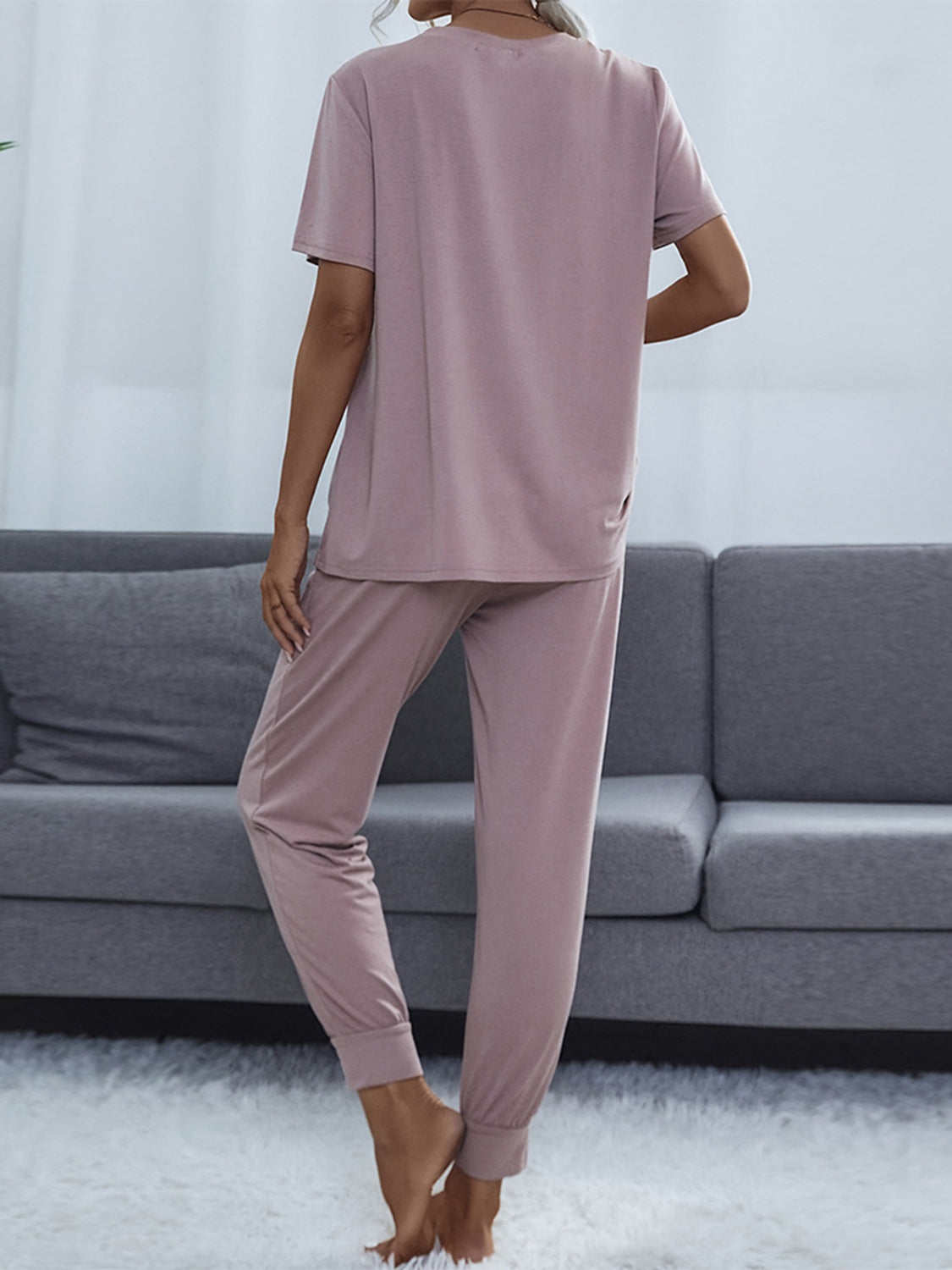 NEWYORKLISTED Intimate Apparel: Person wearing a mauve pajama set standing in a living room with a gray sofa.