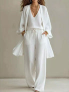 NEWYORKLISTED STYLE COLLECTION: A woman modeling a white three-piece linen outfit consisting of a V-neck top, matching wide-leg pants with a drawstring, and a lightweight open-front cardigan. She is accessorized with layered gold necklaces and bracelets, standing against a neutral beige studio background.