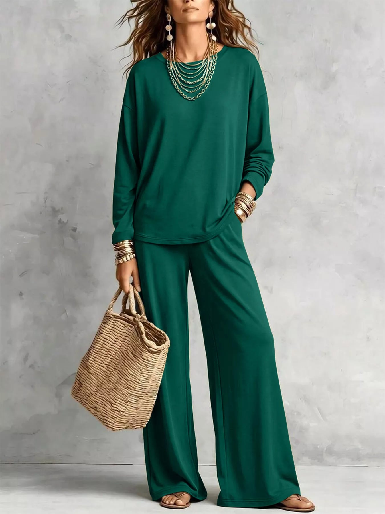 NEWYORKLISTED STYLE COLLECTION: A woman models a matching dark green long-sleeve top and wide-leg pants set, styled with layered gold necklaces, bracelets, and a woven straw tote bag against a textured gray studio backdrop.