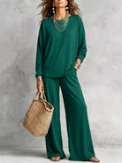 NEWYORKLISTED STYLE COLLECTION: A woman models a matching dark green long-sleeve top and wide-leg pants set, styled with layered gold necklaces, bracelets, and a woven straw tote bag against a textured gray studio backdrop.