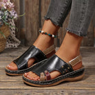 NEWYORKLISTED SHOE COLLECTION: A pair of women's black leather platform slide sandals with cutout toes, beige ankle straps, and decorative gold-tone studs, shown on a model's feet on a rustic wooden floor.