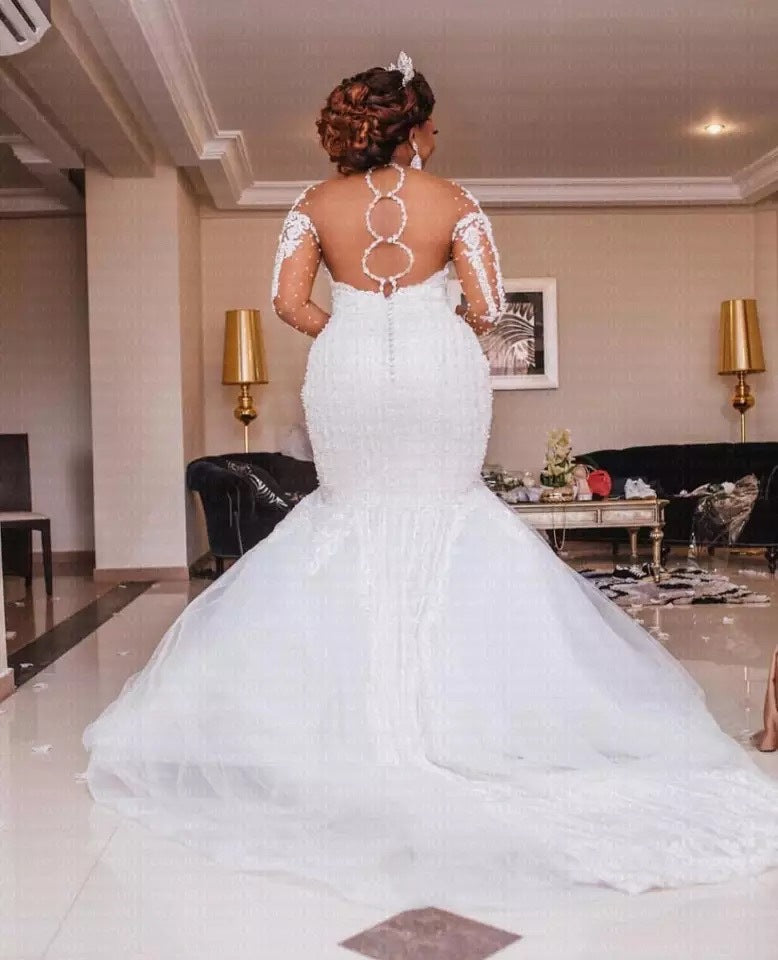 Woman in a white mermaid-style wedding dress with lace sleeves in an indoor setting.