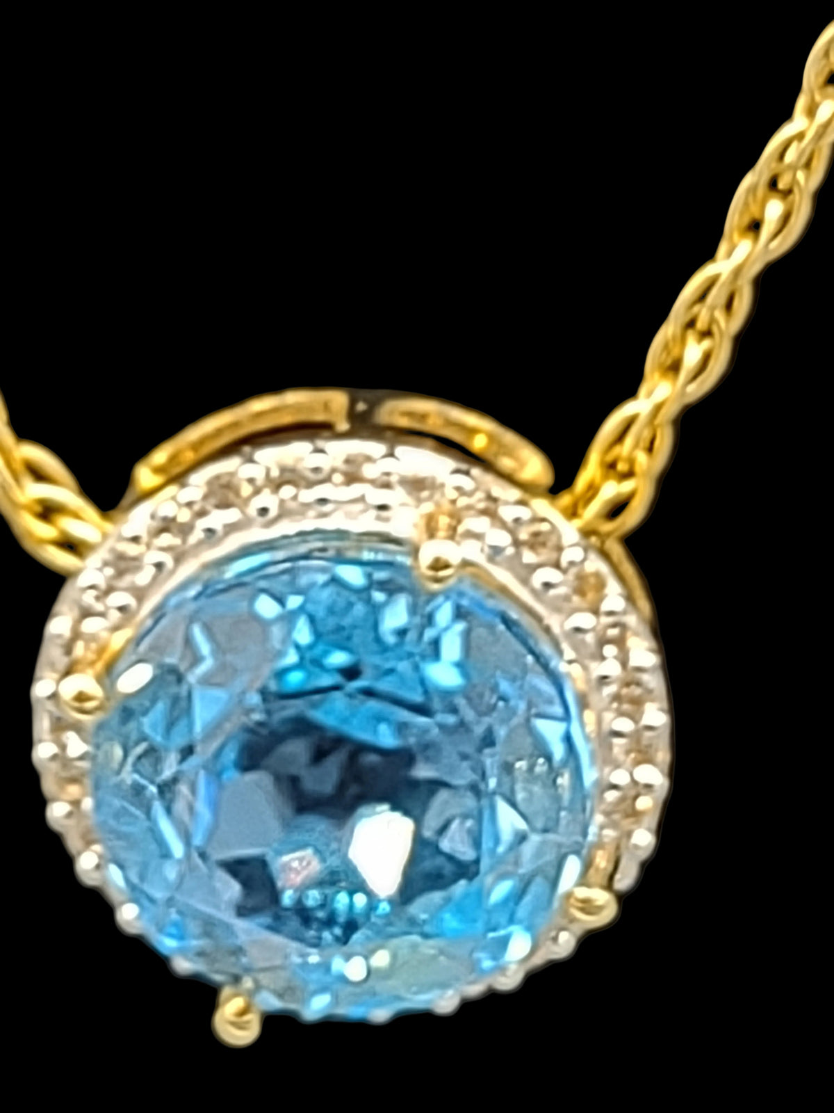 NEWYORKLISTED VINTAGE JEWELRY COLLECTION: A close-up of a gold pendant necklace featuring a large round light blue gemstone surrounded by a halo of small clear stones, set against a black background.