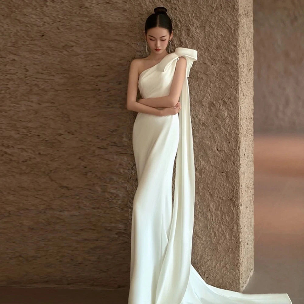 Woman in a white one-shoulder dress standing against a textured brown wall.