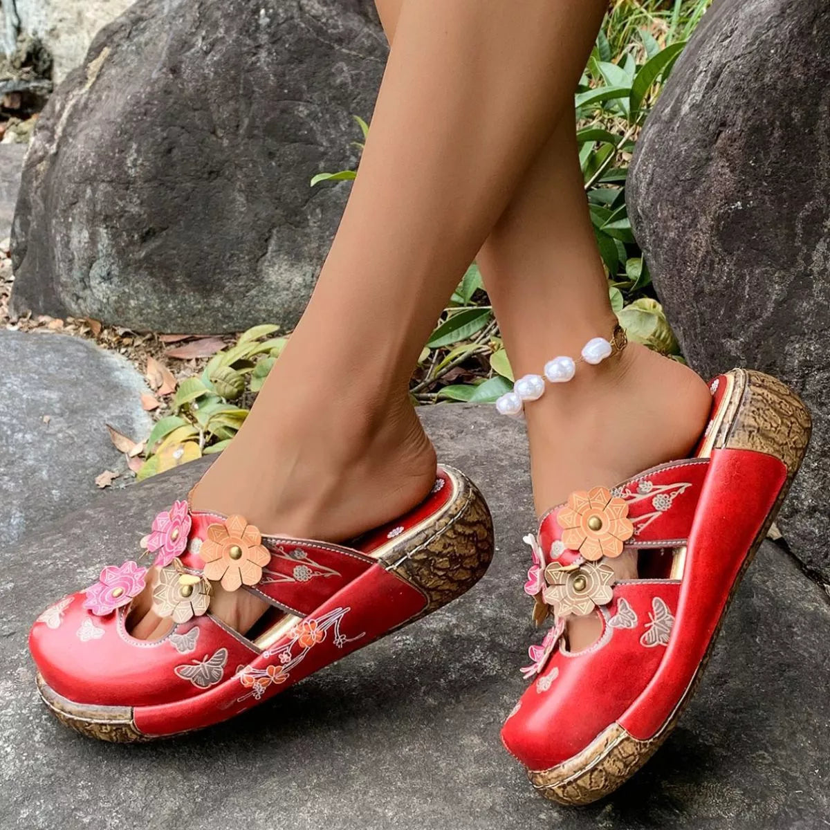 NEWYORKLISTED SHOE COLLECTION: Red floral sandals worn by a person on a stone surface with rocks and grass in the background.