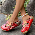 NEWYORKLISTED SHOE COLLECTION: Red floral sandals worn by a person on a stone surface with rocks and grass in the background.