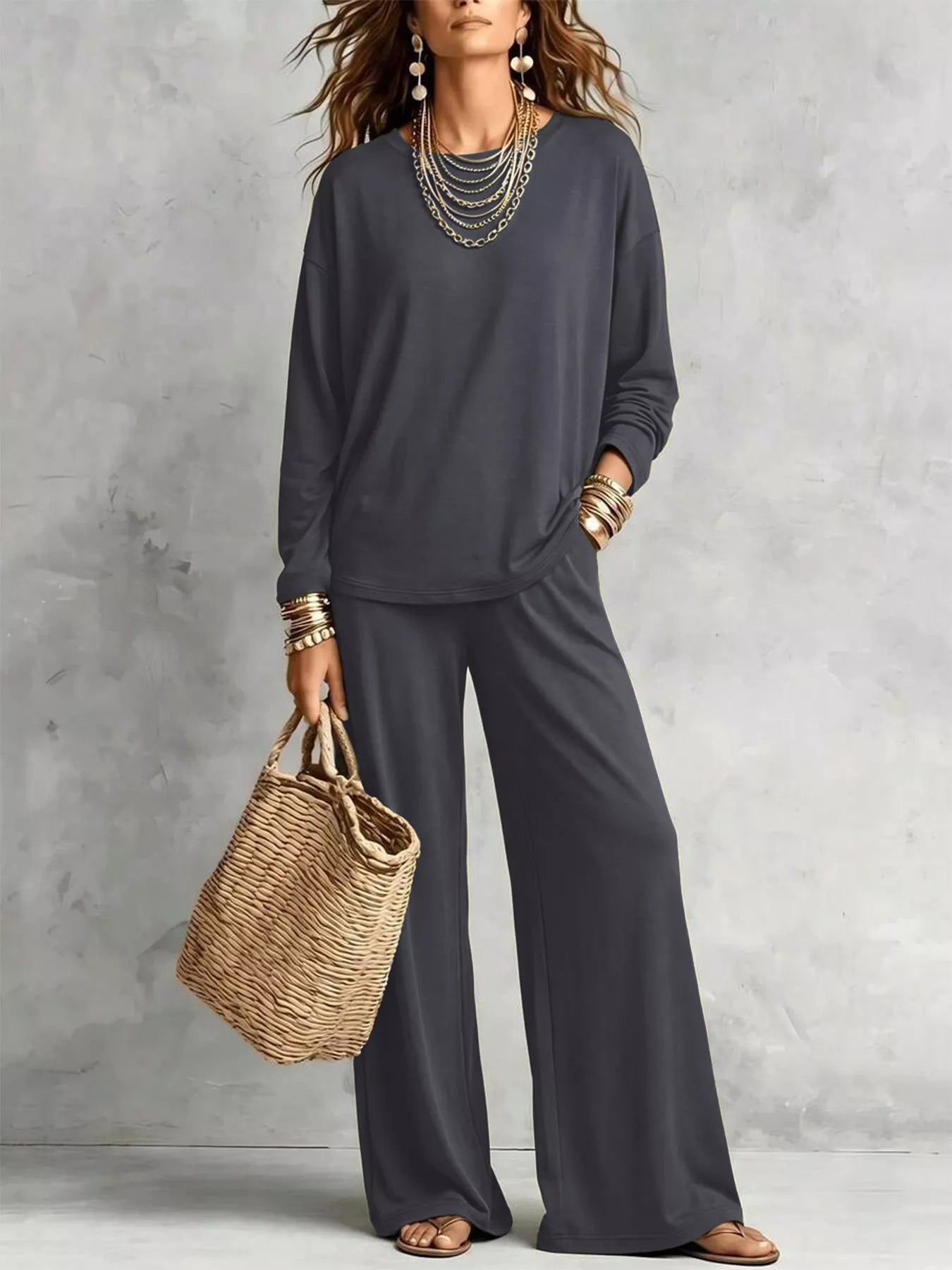 NEWYORKLISTED STYLE COLLECTION: A woman models a dark gray two-piece lounge set consisting of a long-sleeve top and matching wide-leg pants, styled with layered gold necklaces, bracelets, earrings, and a straw tote bag against a textured gray wall.