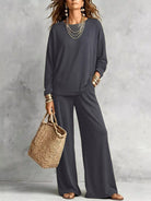 NEWYORKLISTED STYLE COLLECTION: A woman models a dark gray two-piece lounge set consisting of a long-sleeve top and matching wide-leg pants, styled with layered gold necklaces, bracelets, earrings, and a straw tote bag against a textured gray wall.