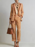 NEWYORKLISTED STYLE COLLECTION: A model wears a matching orange and beige checkered blazer and cropped trousers suit with brown leather trim, styled with a cream shirt, sunglasses, a brown leather handbag, and brown strappy heels against a light gray studio background.