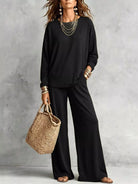 NEWYORKLISTED STYLE COLLECTION: A woman models a black long-sleeve top and matching wide-leg pants, styled with layered gold necklaces, bracelets, and a woven straw tote bag against a textured gray wall.
