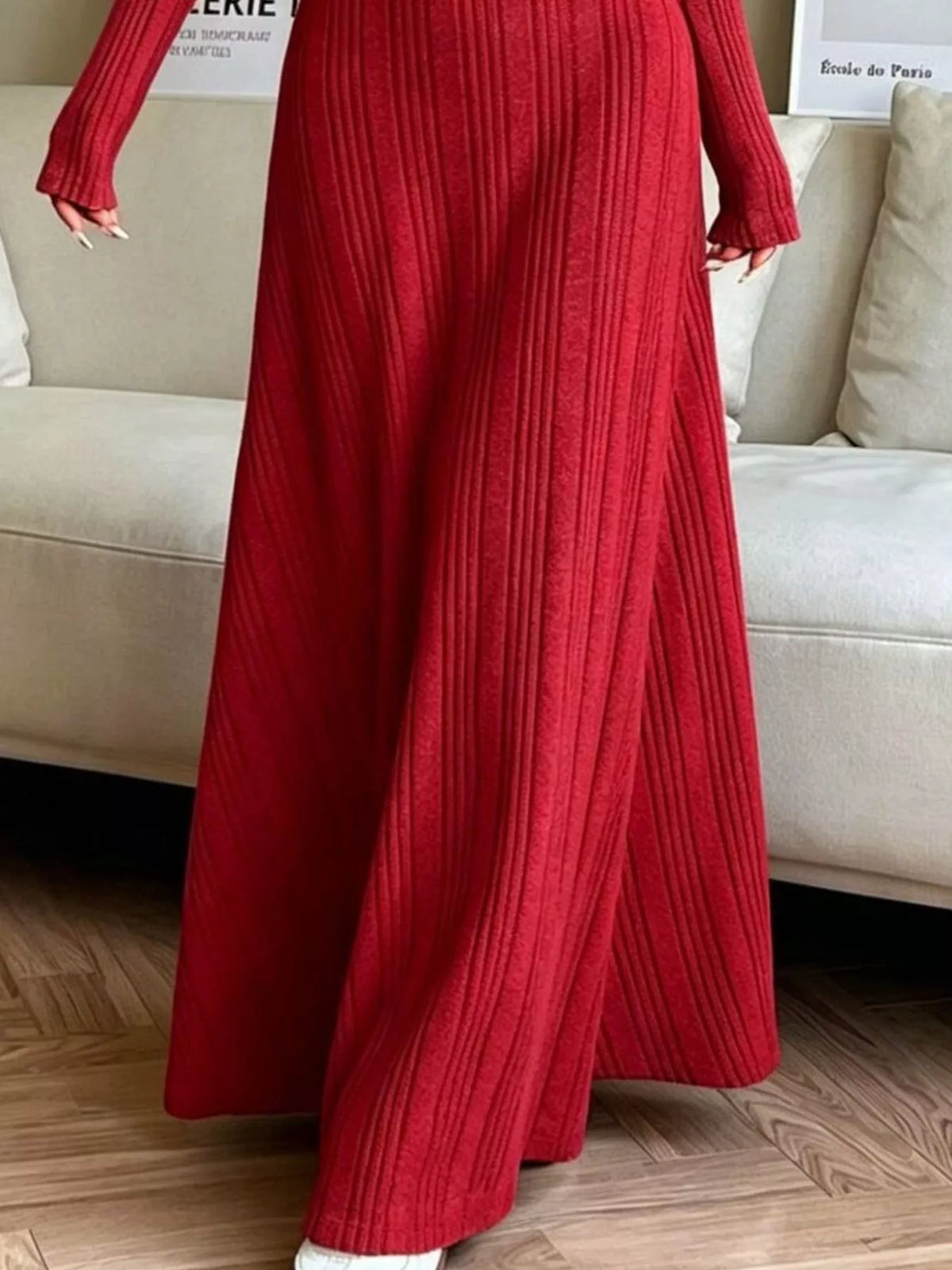 NEWYORKLISTED STYLE COLLECTION: A person wearing a long-sleeve, floor-length red ribbed knit dress standing in front of a light beige sofa on a wood floor.