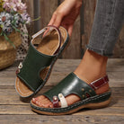 NEWYORKLISTED SHOE COLLECTION: A person wearing a pair of dark green leather slide sandals with burgundy straps and white contrast stitching, standing on a rustic wooden floor while holding the other sandal.