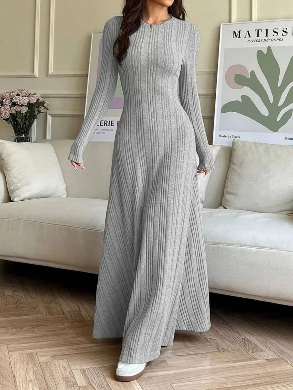 NEWYORKLISTED STYLE COLLECTION: A woman wearing a long-sleeve, ribbed knit maxi dress in light gray, standing in a living room setting with a beige sofa and framed wall art in the background.