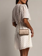 NEWYORKLISTED STYLE COLLECTION: Woman wearing a white lace dress holding a beige handbag against a gray background