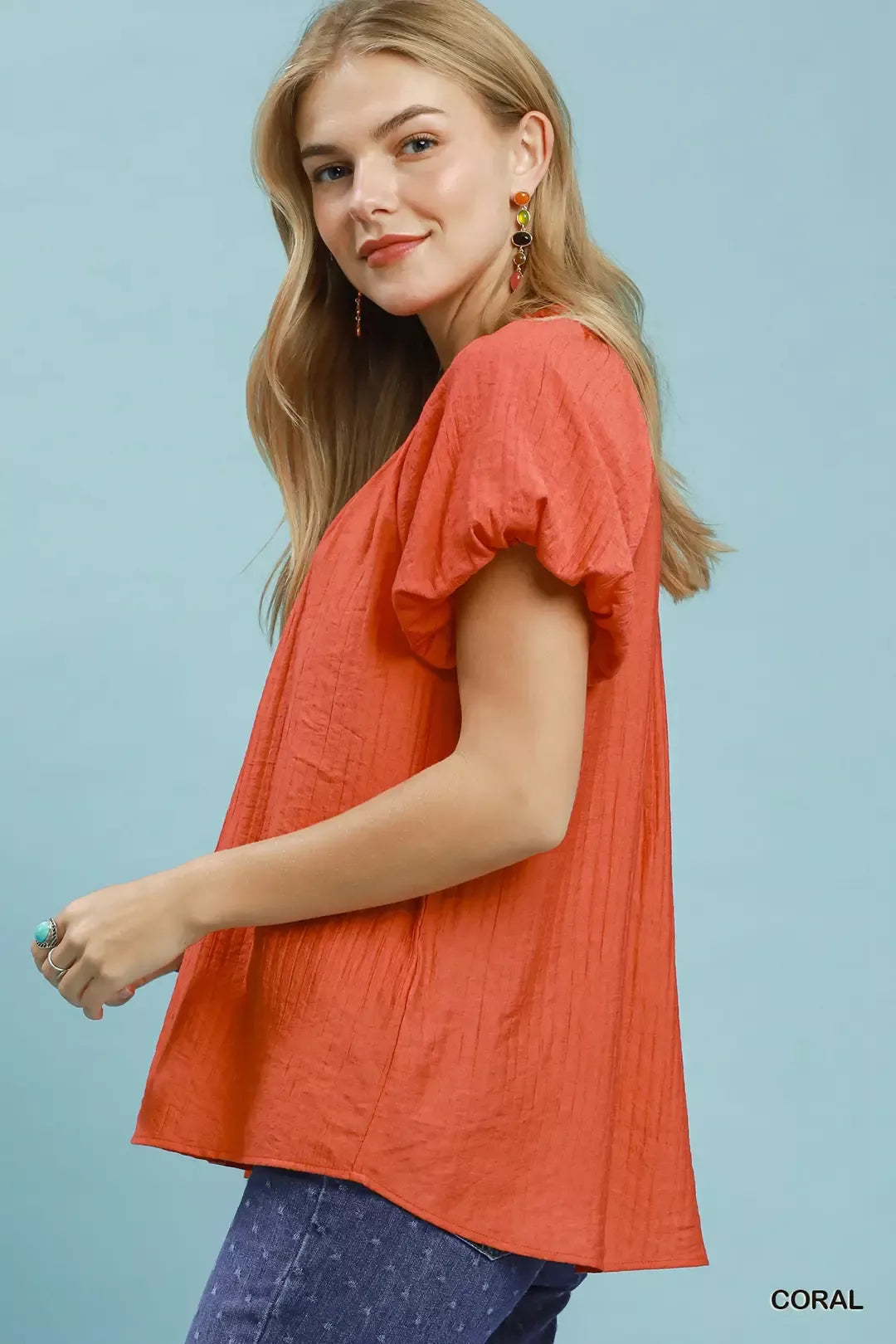 NEWYORKLISTED STYLE COLLECTION: Woman wearing a coral top with ruffled sleeves against a light blue background