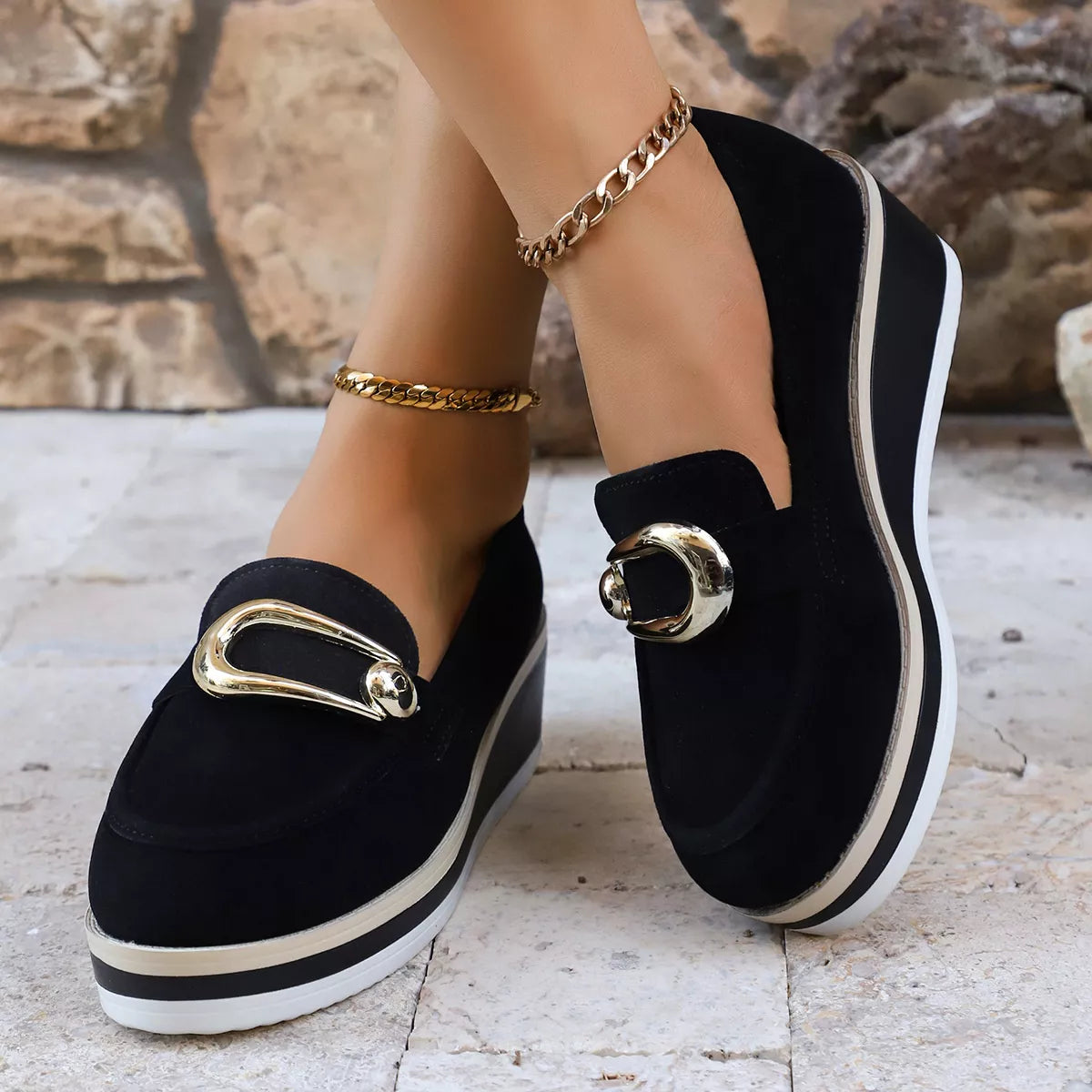 NEWYORKLISTED SHOE COLLECTION: Black loafers with gold buckles worn by a person against a stone wall background