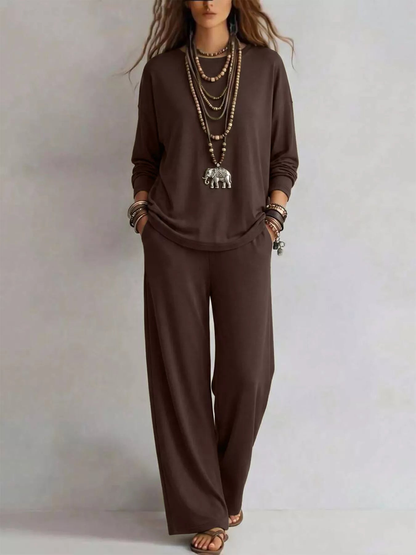 NEWYORKLISTED STYLE COLLECTION: A woman modeling a dark brown long-sleeve top and matching wide-leg pants set, accessorized with layered necklaces, bracelets, and sandals, against a neutral studio background.
