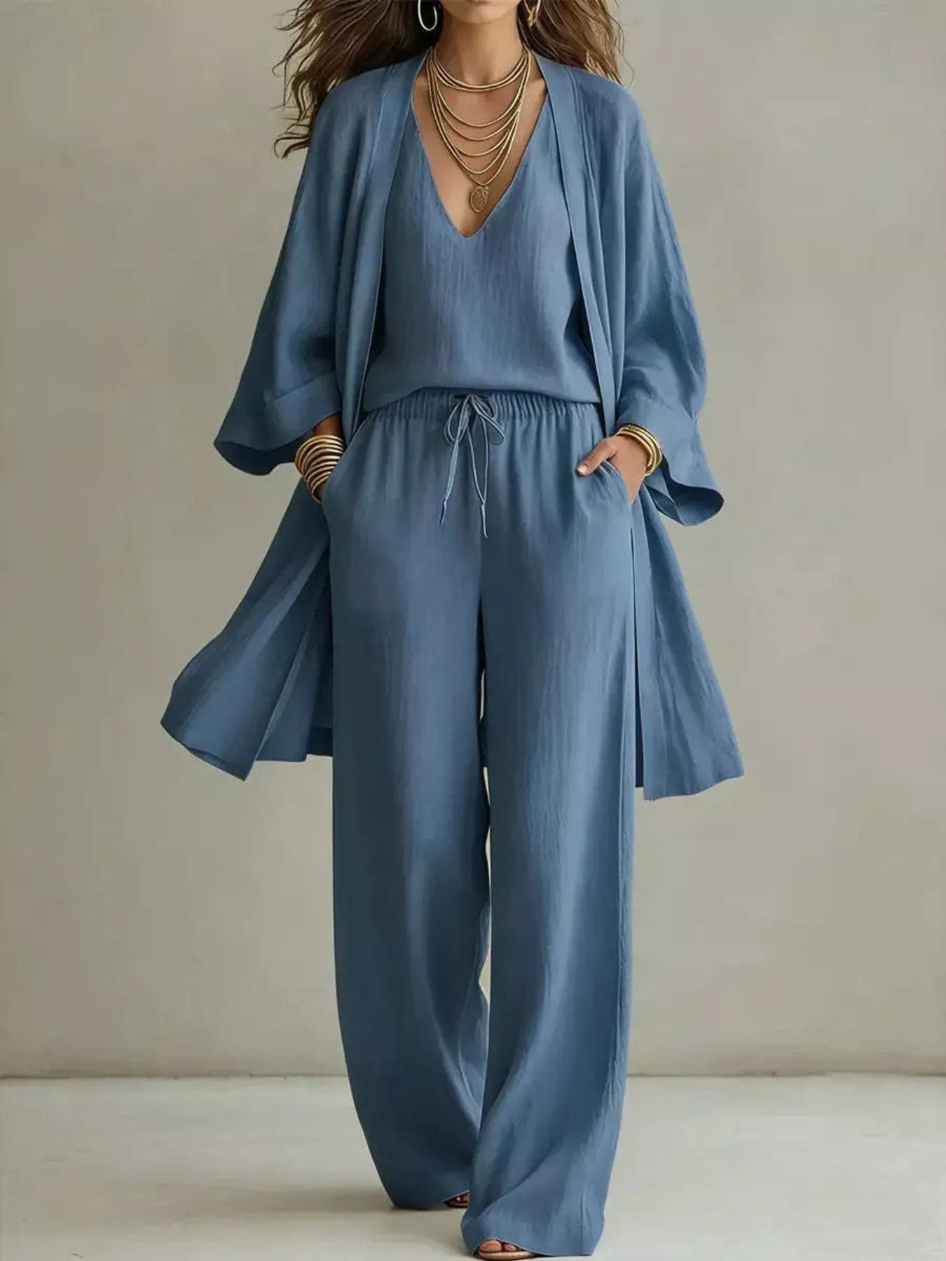 NEWYORKLISTED STYLE COLLECTION: A woman models a dusty blue linen-blend jumpsuit with a matching open-front cardigan. The jumpsuit features a V-neck, wide sleeves, a drawstring waist, and wide-leg pants.
