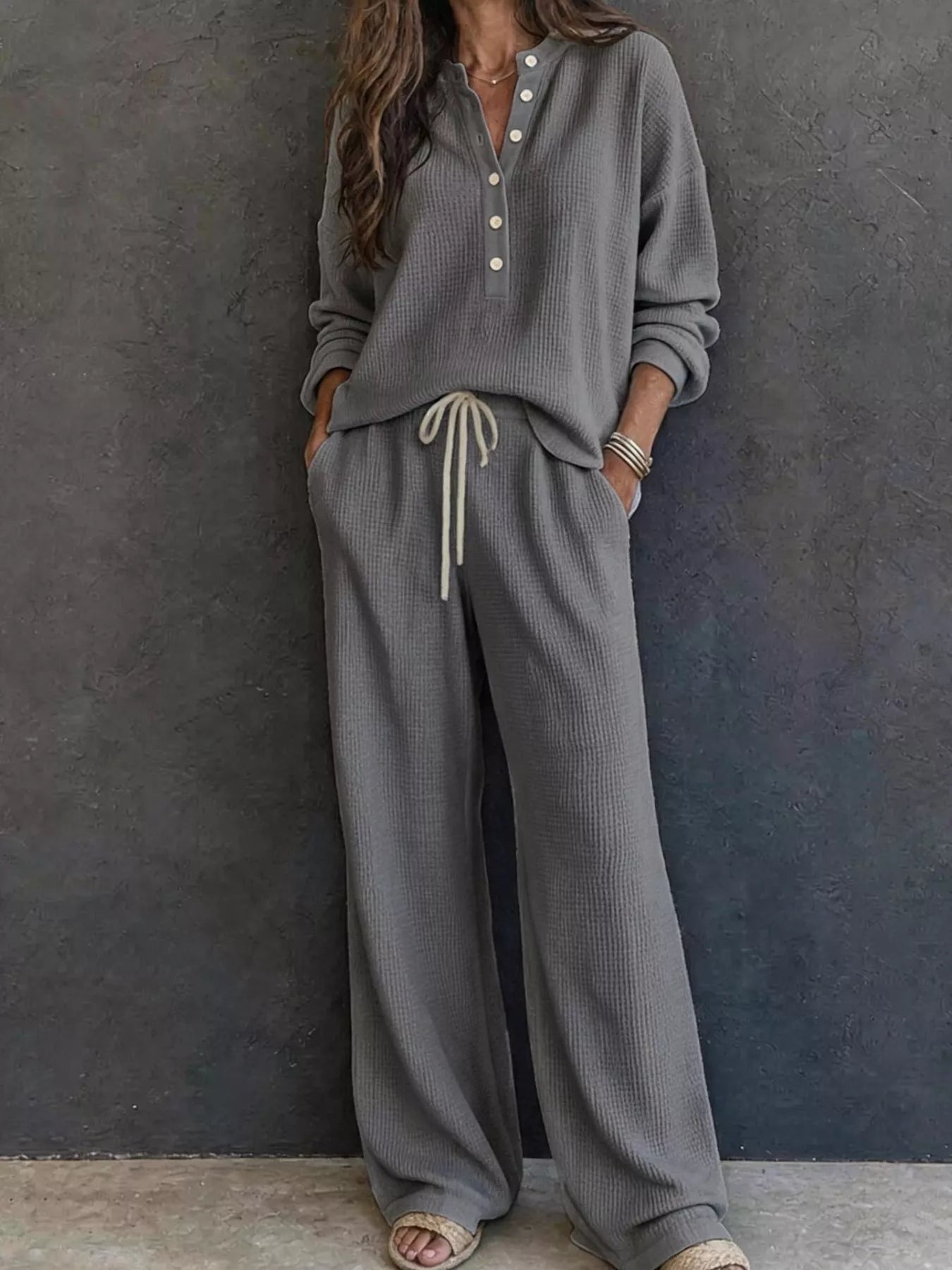NEWYORKLISTED STYLE & COMFORT COLLECTION: A woman wearing a matching gray waffle-knit lounge set with a long-sleeve henley top and wide-leg pants with a drawstring waist, standing against a dark textured wall.