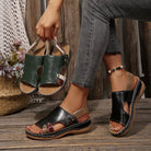 NEWYORKLISTED SHOE COLLECTION: A person wearing black and green cutout platform sandals with metallic buckle straps, seated on a rustic wooden bench while holding a pair of the sandals. Gray jeans are visible, along with a decorative woven basket and flowers in the background.