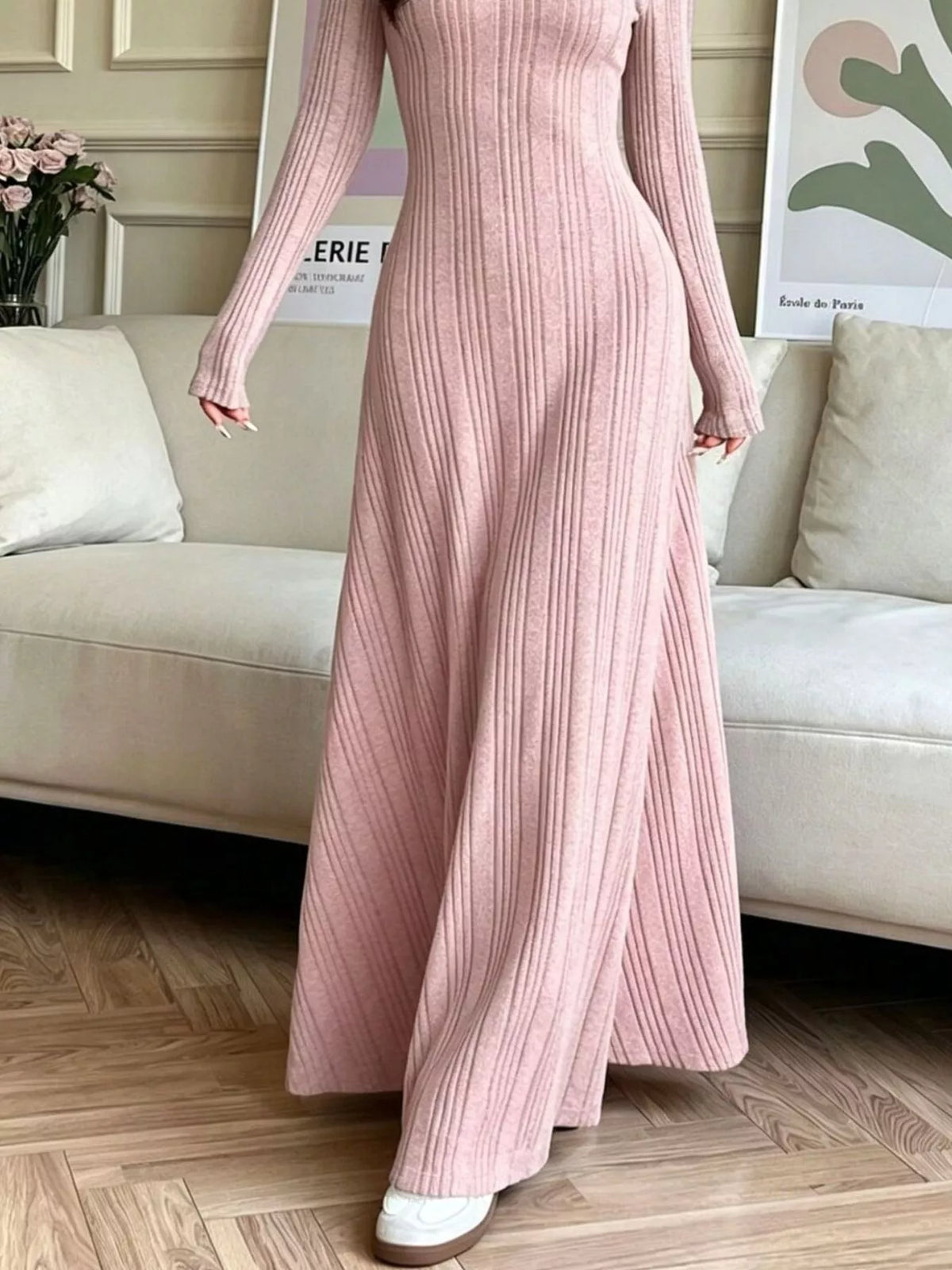 NEWYORKLISTED STYLE COLLECTION: A woman wearing a long-sleeve, ribbed knit maxi dress in a dusty pink color, standing in a living room setting with a beige sofa and wood flooring.