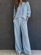 NEWYORKLISTED STYLE & COMFORT COLLECTION: A woman wearing a light blue ribbed knit two-piece loungewear set with a long-sleeve henley top and matching wide-leg pants featuring a white drawstring waist, standing against a dark textured wall.