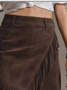NEWYORKLISTED STYLE COLLECTION: A close-up of a dark brown suede skirt with diagonal fringe detailing, worn by a model in a cropped white top.