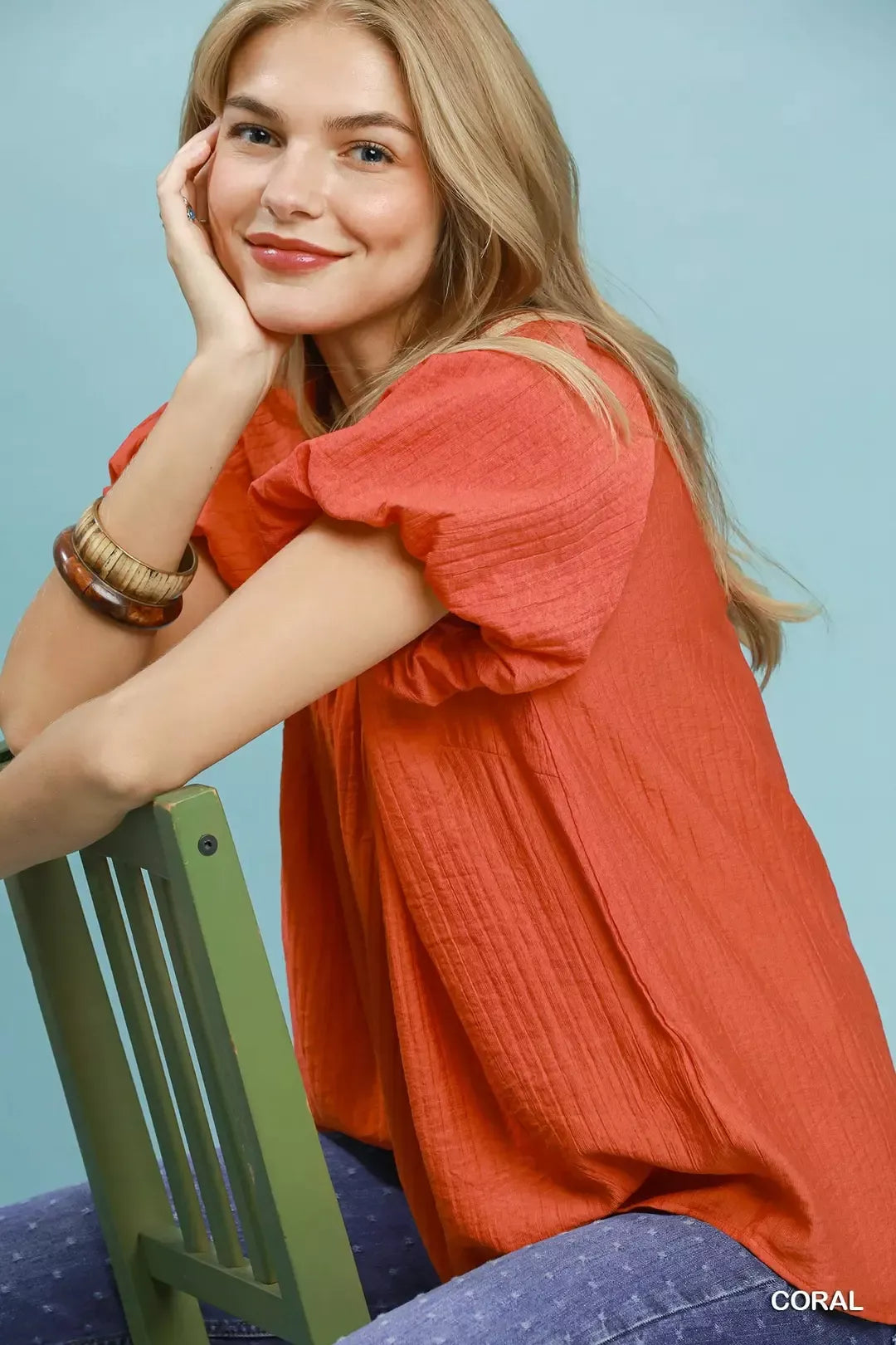 NEWYORKLISTED STYLE COLLECTION: Woman wearing a coral shirt sitting on a green chair against a light blue background