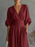 NEWYORKLISTED STYLE COLLECTION: A woman wearing a deep burgundy wrap-style dress with a V-neckline, gathered waist, and elbow-length puff sleeves, standing against a soft gray studio background.