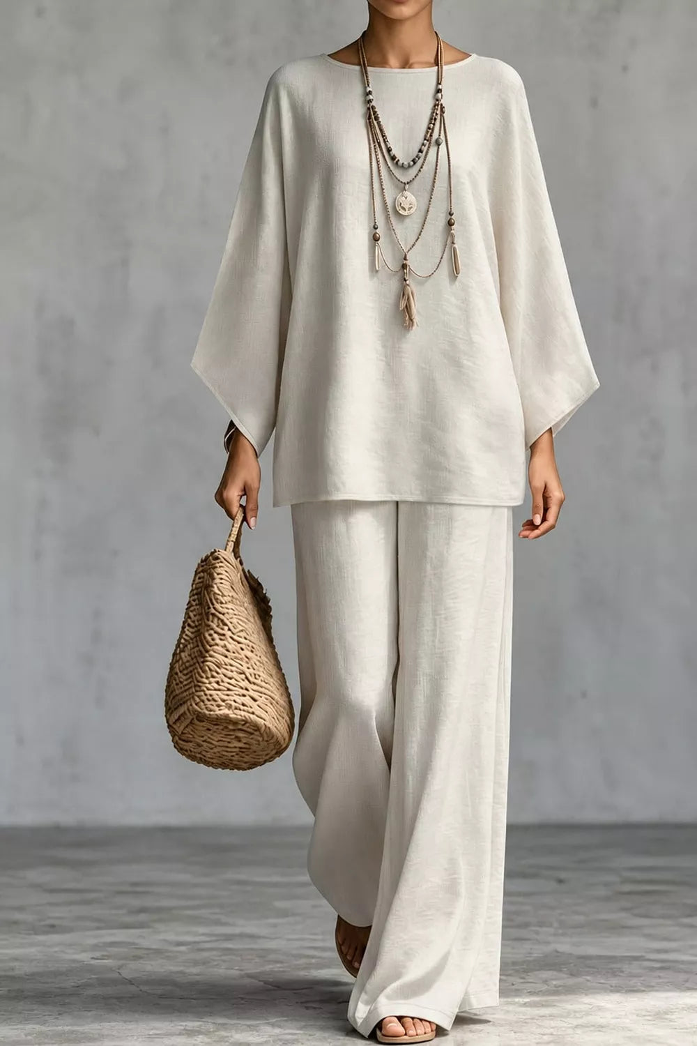 NEWYORKLISTED STYLE COLLECTION: A model wearing a cream-colored two-piece linen outfit consisting of a loose-fitting short-sleeve top and matching wide-leg pants, accessorized with layered necklaces and holding a woven straw bag against a gray concrete background.