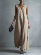 NEWYORKLISTED STYLE COLLECTION: A woman models a sleeveless, V-neck maxi dress in a light beige linen-like fabric, holding a small black clutch against a textured gray studio background.