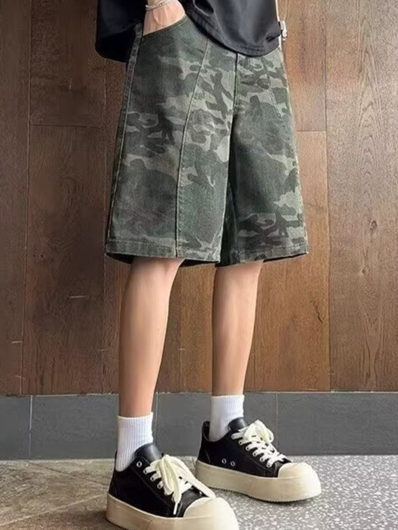 NEWYORKLISTED ATLAS MEN's COLLECTION: Person wearing a camouflage skirt with black sneakers and white socks against a wooden wall.