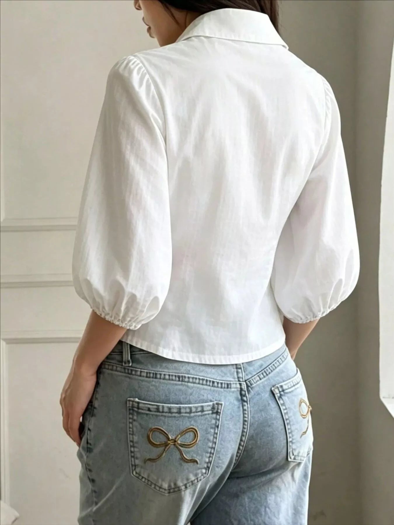NEWYORKLISTED STYLE COLLECTION: A woman shown from the back wearing a white puff-sleeve collared blouse tucked into light-wash blue jeans with decorative bow embroidery on the back pockets.