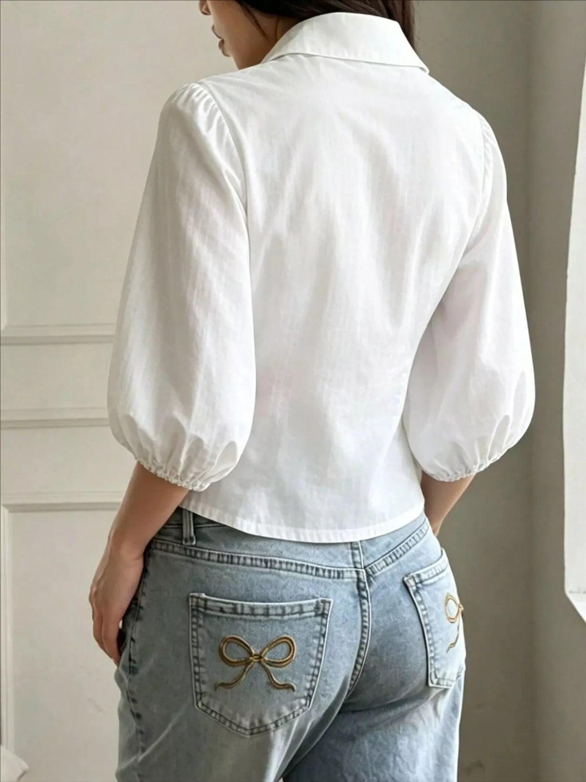NEWYORKLISTED STYLE COLLECTION: A woman shown from the back wearing a white puff-sleeve collared blouse tucked into light-wash blue jeans with decorative bow embroidery on the back pockets.
