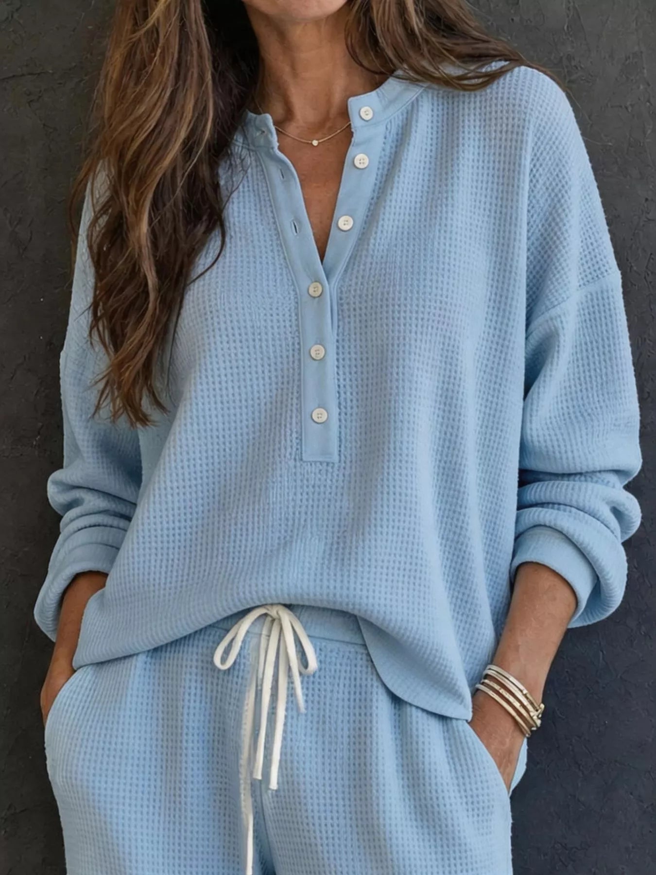 NEWYORKLISTED STYLE & COMFORT COLLECTION: A woman wearing a light blue waffle-knit long-sleeve henley top with white buttons and matching light blue waffle-knit pants with a white drawstring, posed against a dark textured wall.