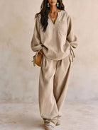 NEWYORKLISTED STYLE COLLECTION: A woman models a beige linen two-piece lounge set consisting of a long-sleeve V-neck top with tie details at the cuffs and matching wide-leg trousers, set against a textured beige wall.