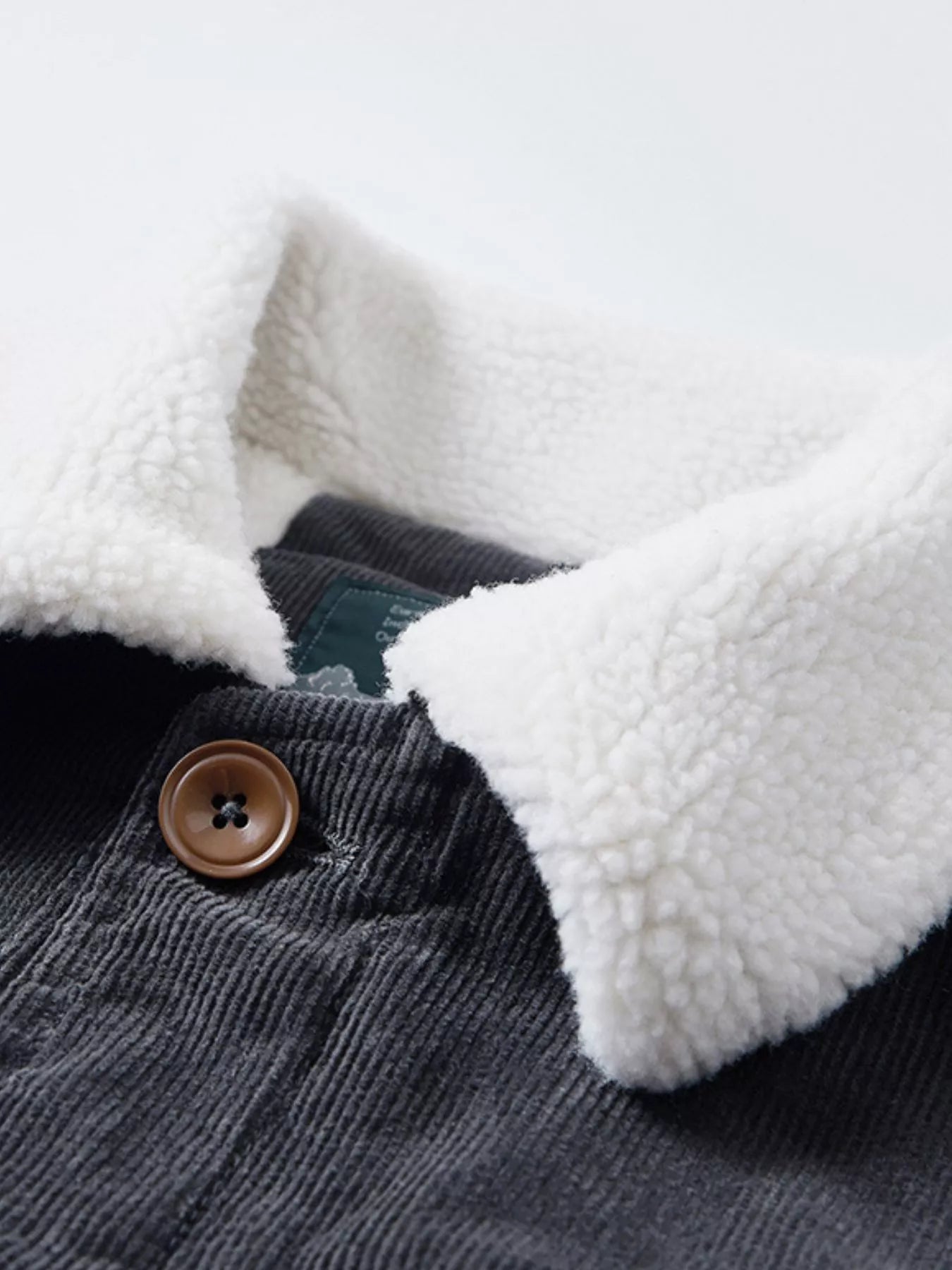 NEWYORKLISTED ATLAS MEN'S COLLECTION: Close-up of a gray corduroy jacket with a white sherpa lining and a button, on a light gray background.