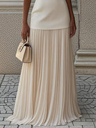 NEWYORKLISTED STYLE COLLECTION: Person wearing a beige pleated skirt holding a matching handbag on a stone pavement.