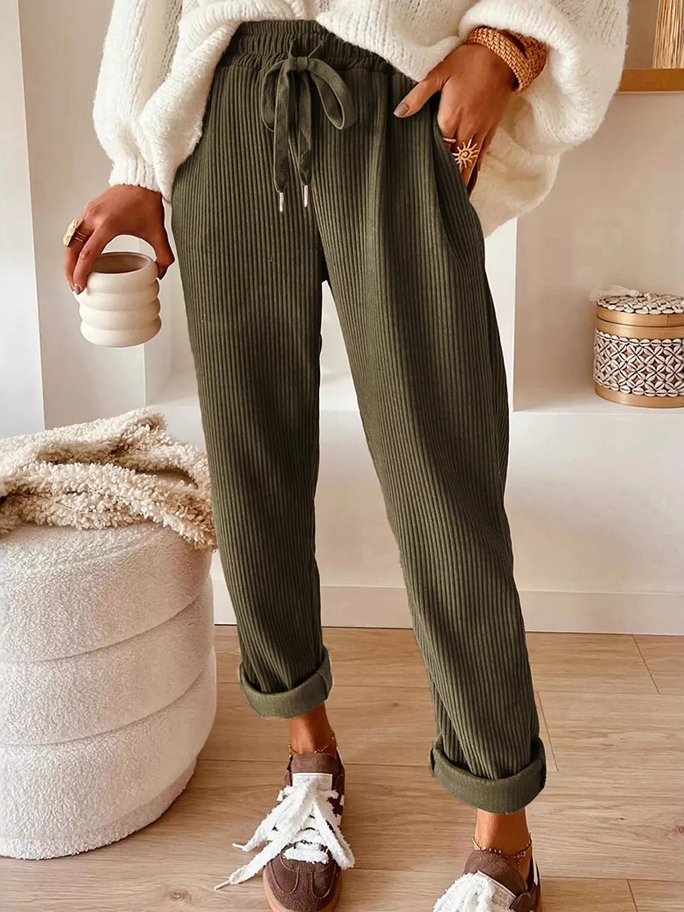 NEWYORKLISTED STYLE COLLECTION: Person wearing green ribbed pants with a white sweater and brown shoes, holding a cup.