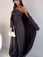 NEWYORKLISTED COMFORT COLLECTION: Woman wearing a dark gray strapless jumpsuit with a white handbag against a plain background